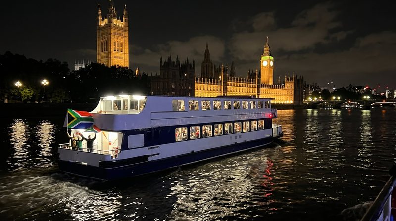 A river boat flying South African flags on the Thames at night, with the Houses of Parliament and Big Ben lit up behind